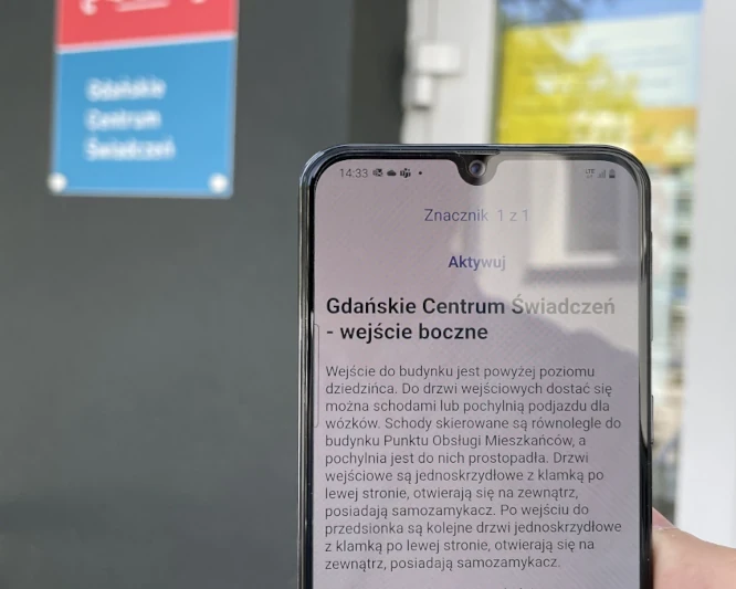 Osoba trzyma smartfon z informacjami o bocznym wejściu do Gdańskiego Centrum Świadczeń, dostarczonymi przez system Totupoint.